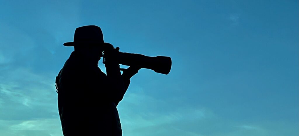 Thomas Reed silhouetted against the early morning sky looking through a camera and telephoto lens